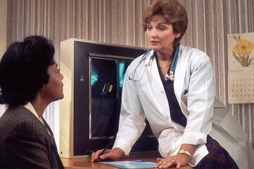 A middle aged white female doctor in a white lab coat talking to a black female lupus patient about a lupus diagnosis